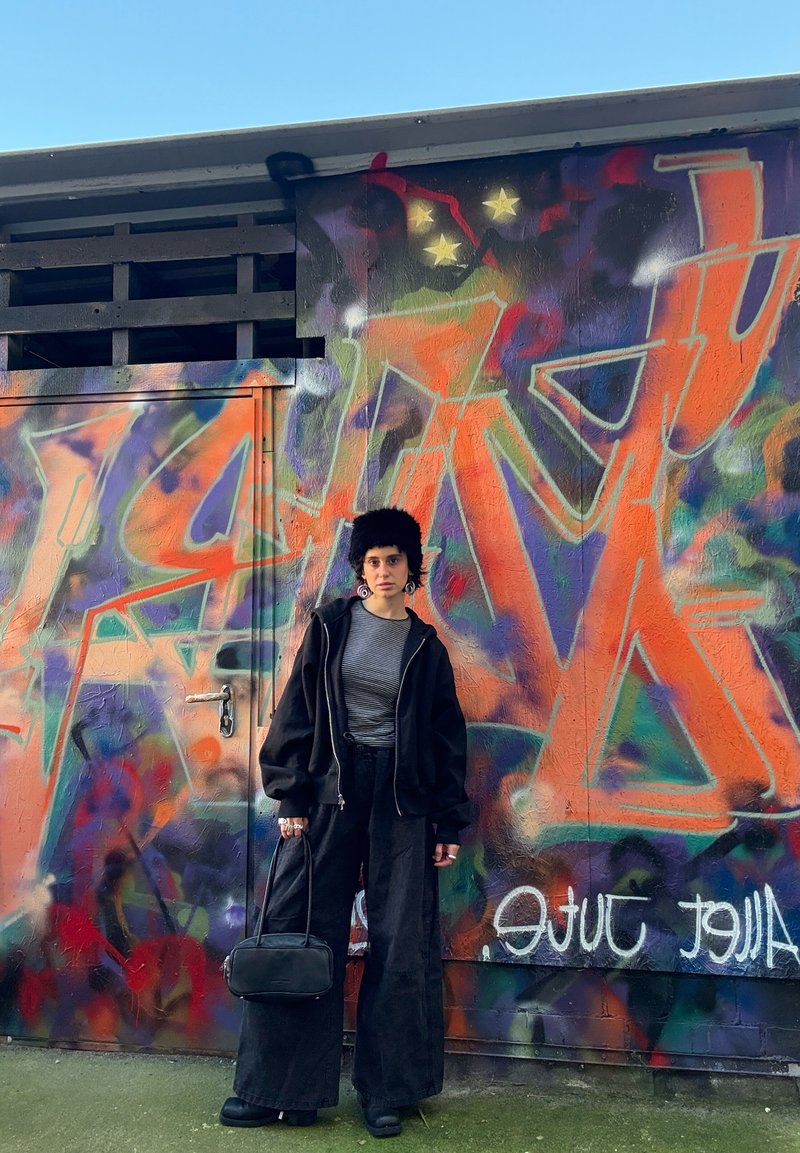 Sweatshirt preta oversized, blusa cinzenta às riscas, calças largas pretas, mala preta, sapatos pretos robustos; fundo com graffiti colorido.