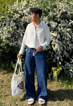 Camisa de lino blanca con botones, pantalones de mezclilla anchos de color azul, sandalias blancas y un bolso de mano beige con un logo. Fondo de follaje verde y flores blancas.