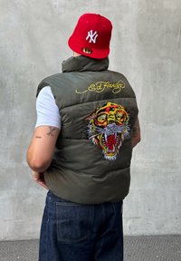 Gilet imbottito verde con un grande ricamo di una tigre e il logo "Ed Hardy" sul retro. Abbinato a un cappellino rosso e jeans.
