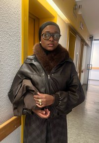 Hnědá kožená bomber bunda s kožešinovým límcem, nošená přes černý top, s výraznými brýlemi a zlatým prstenem na ruce.