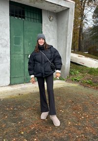 Černá prošívaná bunda s bílými manžetami, černé kalhoty a šedé boty. Stojí před zelenými dveřmi, má šedou čepici a malou tašku.