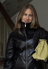 Młoda kobieta z długimi blond włosami, ubrana w czarną skórzaną kurtkę na zamek, trzyma żółtą torebkę ze sznurkiem wewnątrz pomieszczenia.