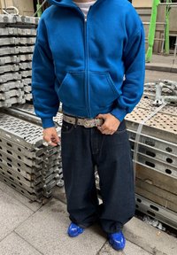 Personne portant un sweat à capuche zippé bleu, un jean ample foncé, une ceinture à motifs, plusieurs bagues et des baskets bleues, debout près d'un échafaudage métallique empilé.