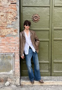 Veste en cuir marron sur une chemise blanche, jean évasé bleu, et bottes pointues marron. Se tenant contre une porte vert olive avec des détails floraux.