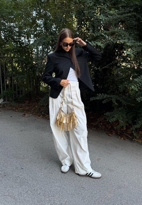 Femme en veste noire, pantalon blanc large et baskets ajustant ses lunettes de soleil, tenant un sac à main à franges dorées, debout sur un chemin près de buissons verts.