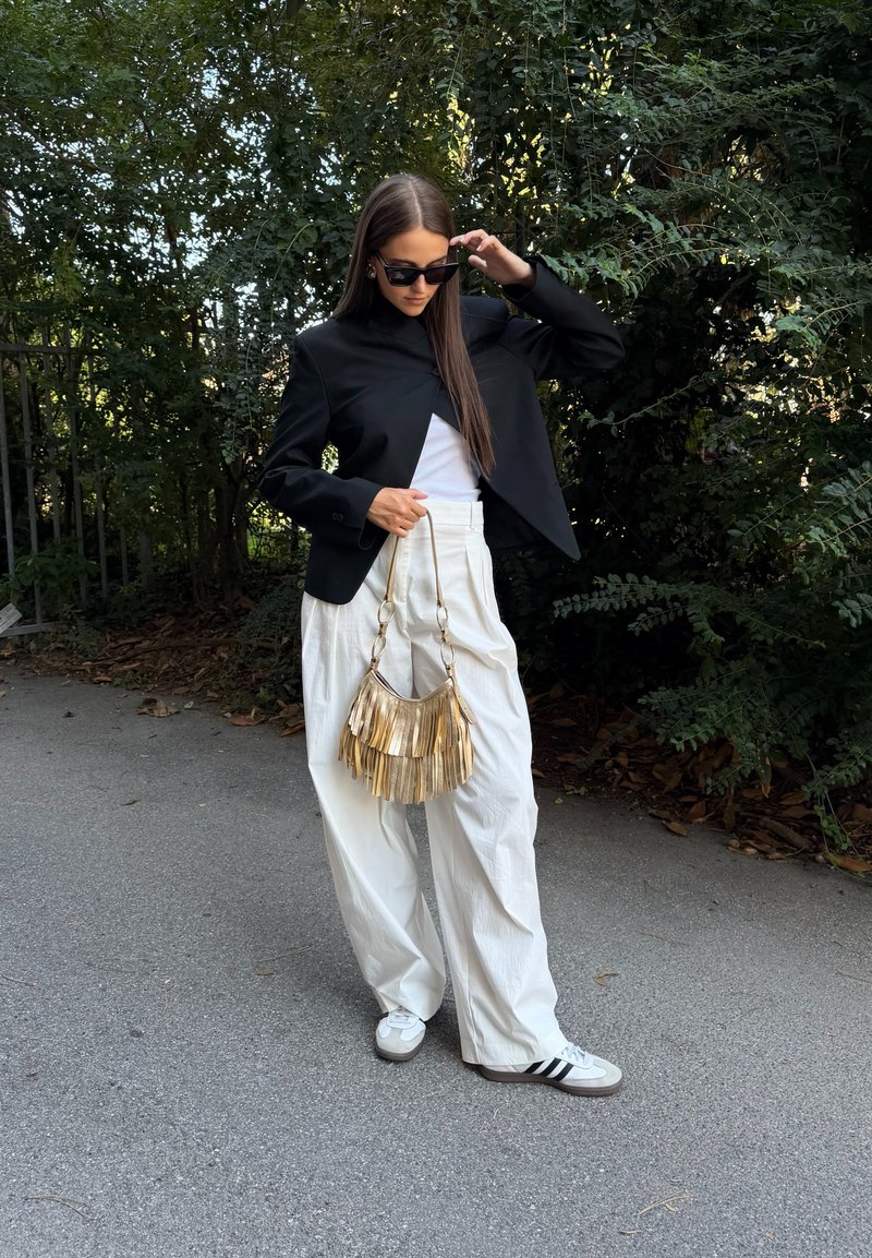 Donna con giacca nera, pantaloni bianchi ampi e sneakers che si sistema gli occhiali da sole, tenendo una borsa con frange dorate, in piedi su un sentiero vicino a cespugli verdi.