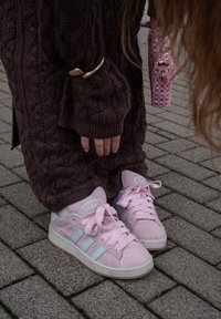 Baskets en daim rose avec des rayures, des lacets et un logo bleu clair. Portées avec un pull oversize marron texturé et un pantalon à motifs.
