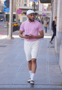 Polo rosa a maniche corte, pantaloni corti bianchi su misura, calze bianche e scarpe bicolore. Cappellino bianco e telefono in mano, mentre cammina su un marciapiede cittadino.