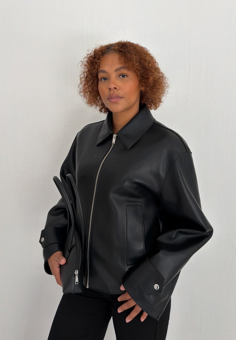Jeune personne aux cheveux bouclés portant une veste en similicuir noire, tenant un sac noir zippé assorti sur un fond clair uni.