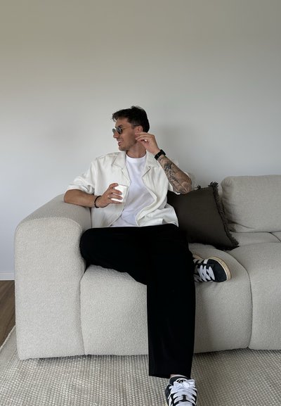 Hombre con gafas de sol, camisa blanca y pantalones negros sentado en un sofá beige sosteniendo una taza blanca, mirando hacia su izquierda con una leve sonrisa.
