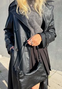 Sort læder trench coat med knapper og brede revers, parret med en grå striktrøje og en plisseret sort nederdel. Holder en lille sort håndtaske.