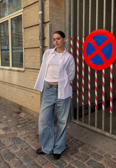 Camisa blanca de botones sobre una camiseta blanca ajustada, combinada con jeans de mezclilla azul de pierna ancha. Mocasos negros, de pie frente a una puerta de rejas.