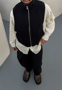 Fekete kötött ujjatlan mellény magas nyakkal és cipzárral, fehér hosszú ujjú ing fölött viselve, laza fekete nadrággal és sötét cipővel párosítva.