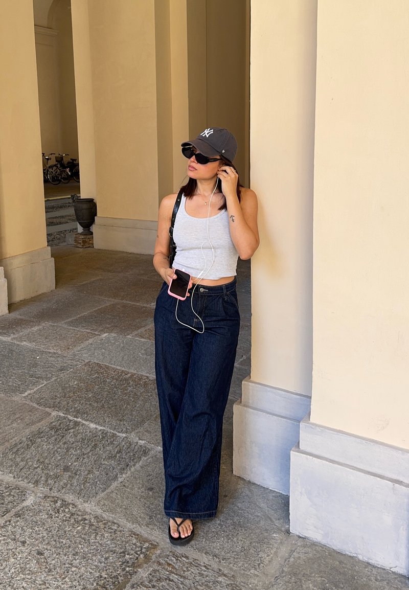 Femme portant des lunettes de soleil et une casquette écoute des écouteurs tout en tenant un smartphone, debout contre une colonne dans un passage couvert extérieur.