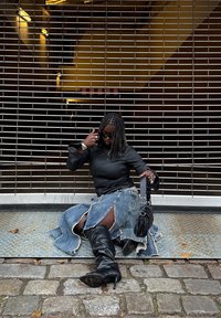 Čierny kožený top s dlhými rukávmi, modrá rifľová sukňa s roztŕhanými okrajmi, čierne čižmy nad kolená a malá čierna kabelka, sediaca opretá o kovový rošt.