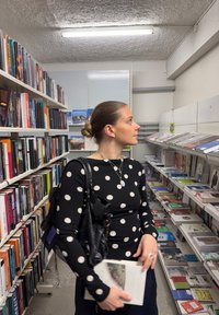 Γυναίκα με μαύρο τοπ με πουά, κρατώντας ένα βιβλίο, περιηγείται σε ράφια γεμάτα βιβλία και περιοδικά σε ένα καλά φωτισμένο διάδρομο βιβλιοπωλείου.