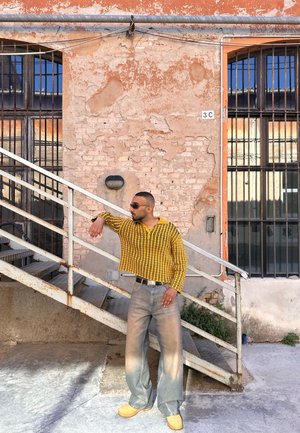 Camisa amarilla a rayas, jeans grises holgados y zuecos en un hombre que está de pie junto a unas escaleras de metal contra una pared de ladrillo desgastada con ventanas.
