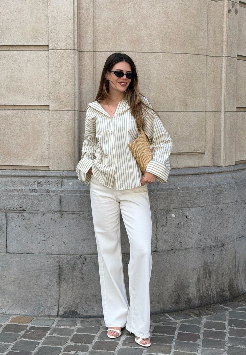 Woman wearing sunglasses, striped button-up shirt, white wide-leg pants, white sandals, and holding a woven clutch beside a stone wall outdoors.