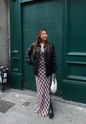 Chaqueta oversize de cuero negro sobre un vestido maxi de cuadros en color burdeos, sosteniendo un bolso blanco con superficie texturizada, en un fondo verde.