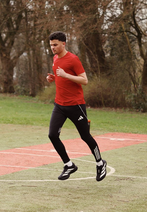 Mladý muž běží na venkovním oválu, má na sobě červené tričko, černé kalhoty Adidas a černé běžecké boty s bílými pruhy.