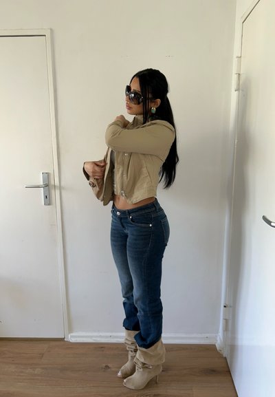 Mujer con chaqueta corta beige, vaqueros azules, botas beige holgadas con tacón y grandes gafas de sol, ajustándose la manga de la chaqueta en el interior junto a puertas blancas.
