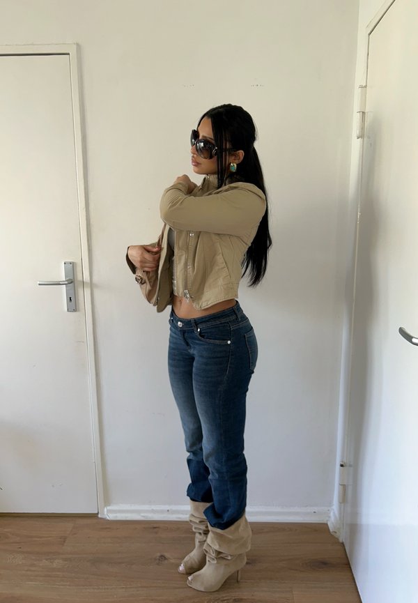 Woman wearing beige cropped jacket, blue jeans, beige slouchy heeled boots, and large sunglasses, adjusting jacket sleeve indoors by white doors.
