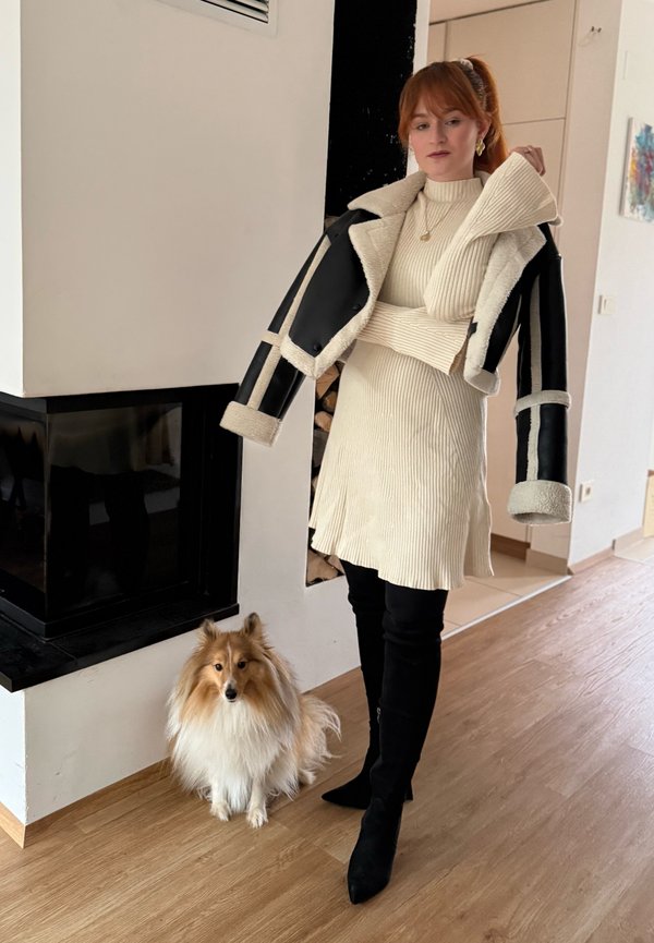 Femme en robe côtelée crème et bottes noires avec une veste en peau retournée noire drapée sur les épaules, debout près d'un Shetland Sheepdog à l'intérieur d'une maison.
