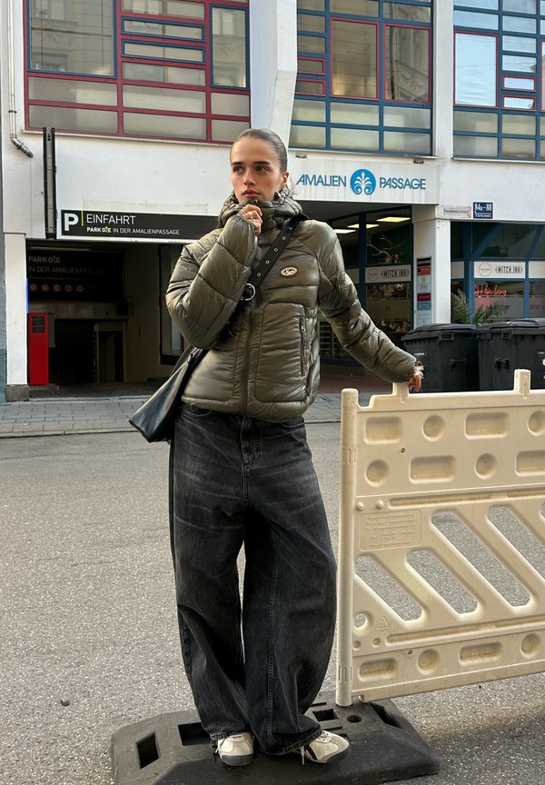 Una mujer con una chaqueta acolchada de color verde oliva y unos vaqueros de pernera ancha está de pie junto a una barrera vial, frente a la entrada de Amalien Passage.