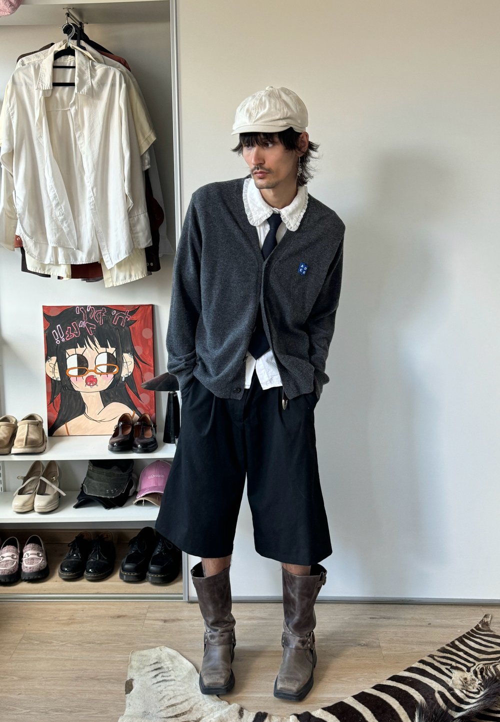 Homme portant une casquette blanche, une chemise à col plissé blanche, un cardigan sombre, un short large noir et des bottes brunes, debout dans une pièce avec des vêtements et des chaussures.