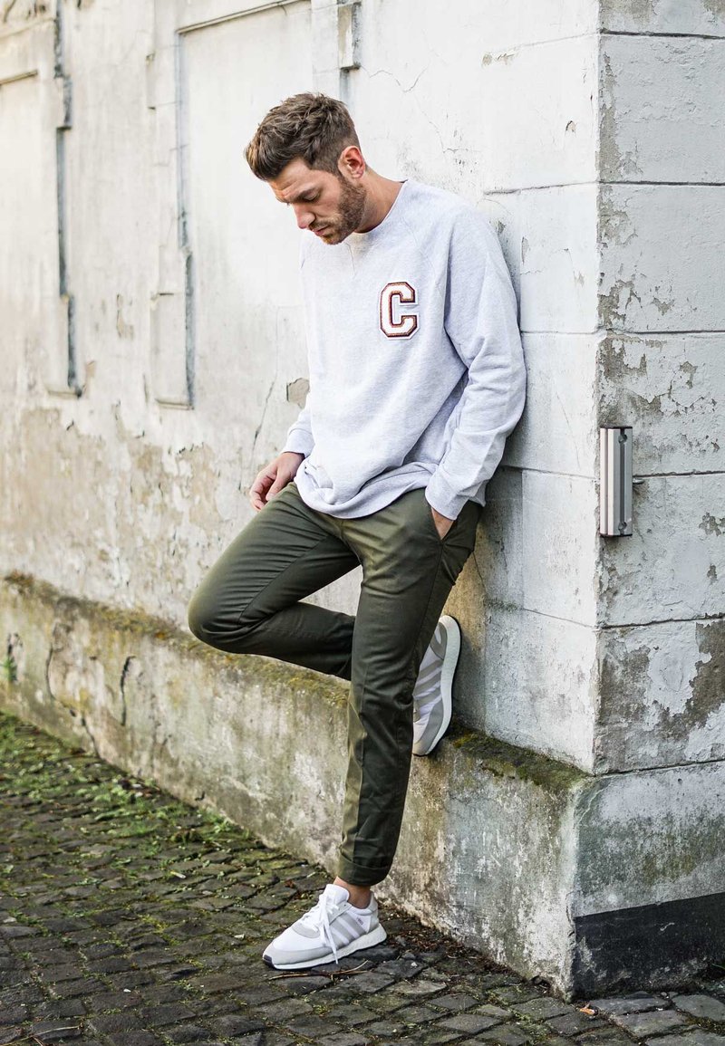 Gray sweatshirt with a "C" logo, olive green slim-fit pants, and white athletic shoes, leaning against a textured wall.
