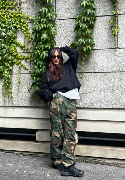 Sudadera negra corta, camisa blanca debajo, pantalones cargo de camuflaje con bolsillos y zapatillas negras. Hiedra verde contra un fondo de pared texturizada.