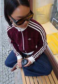 Burgunderfarget kordfløyelsjakke med glidelås og hvite striper, kombinert med mørkeblå vidbente jeans. Holder en svart kaffekopp. Sorte, store solbriller.