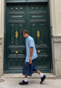 Tânăr cu păr creț, purtând ochelari, o cămașă bleu deschis, fustă din denim, șosete albe și pantofi negri, trecând pe lângă o ușă mare verde ornate.