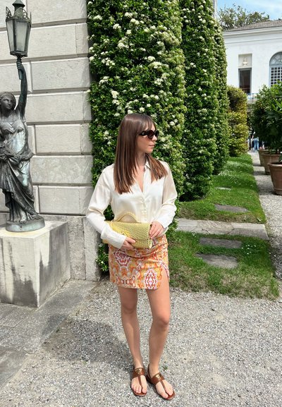 Blusa blanca de seda, minifalda naranja estampada con abertura lateral, bolso amarillo de mimbre, sandalias marrones, rodeada de vegetación y una estatua.