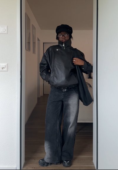 Persona con chaqueta de cuero negra, vaqueros anchos desteñidos, gorra negra, gafas, y llevando un bolso tote negro, de pie en una puerta en el interior.