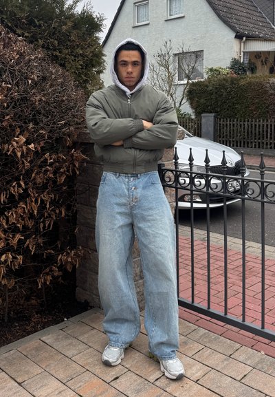 Joven con los brazos cruzados que lleva una chaqueta gris con capucha, vaqueros azul claro de gran tamaño y zapatillas, de pie cerca de una puerta en una zona residencial.