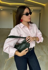 Roza satenasta srajca z gumbi, temno modri džins, zelena pletena torbica, rdeča okrogla uhani, črne sončna očala, gladka notranja postavitev.