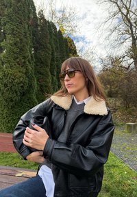 Crna kožna jakna s ovratnikom od ovčje vune, sivi džemper, bijela košulja, drži crnu torbu, na pozadini zelenila i oblačnog neba.