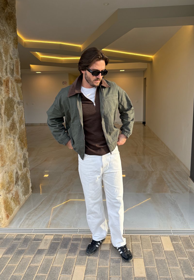 Blouson aviateur vert olive avec col marron, chemise en maille marron, pantalon blanc et baskets noires sur un sol texturé dans un intérieur lumineux.