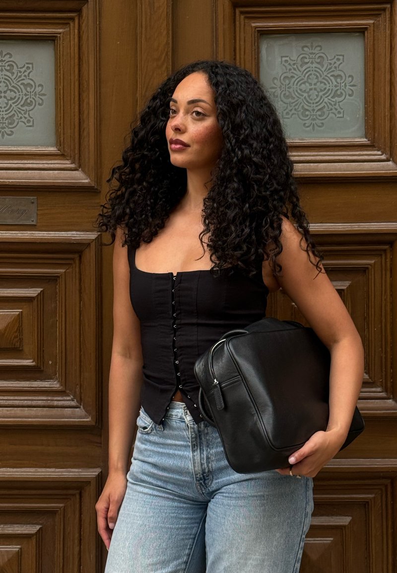 Femme aux longs cheveux bouclés portant un haut noir sans manches et un jean clair, tenant un sac en cuir noir devant une porte en bois.