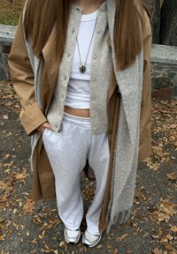 Tânăr purtând pantaloni sport albi, adidași, top scurt alb, cardigan bej, palton maro, eșarfă gri și pandantiv argintiu în aer liber, pe frunze căzute.