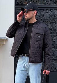 Prošívaná černá bunda, kulatý výstřih, přední zip, kapsa na prsou. K tomu světle modré džíny a černý svetr. Doplňky: černá čepice, sluneční brýle.