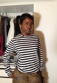 Chemise à manches longues rayée noir et blanc, tissu doux, coupe ajustée ; portée avec un pantalon marron texturé. En arrière-plan, un portant à vêtements.