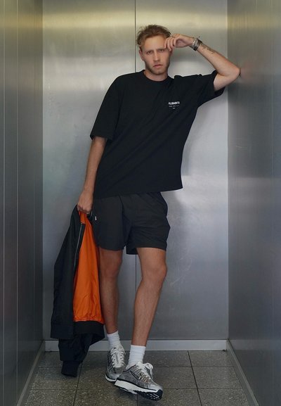 Camiseta negra oversized, pantalones cortos negros y zapatillas plateadas. La chaqueta exterior es negra con un forro naranja. Texturas suaves y diseño sencillo.