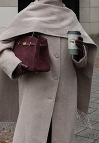 Manteau en laine beige avec grands boutons, châle drapé, sac à main en daim bordeaux avec accents dorés, et une tasse de café verte. Surface texturée, couleurs chaudes.