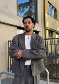 Homme aux cheveux bouclés et lunettes, vêtu d'un costume gris et d'une chemise blanche, debout les bras croisés dans un environnement urbain en plein air.