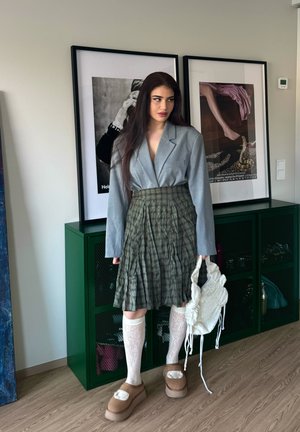 Mujer con cabello largo y oscuro, que lleva un blazer gris, falda verde plisada, calcetines blancos hasta la rodilla y zapatos beige, sosteniendo un bolso blanco texturizado en el interior.