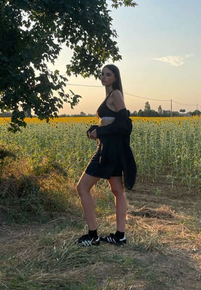 Top corto negro con logotipo blanco, shorts de mezclilla y zapatillas negras con rayas blancas, frente a un campo de girasoles y un fondo de atardecer.