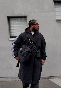 Trench-coat noir avec des rayures blanches, superposé à un sweat à capuche noir. Porte un sac noir avec un logo. Lunettes de soleil foncées, cheveux tressés, devant un mur texturé.