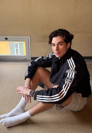 Chaqueta negra de Adidas con rayas blancas, pantalones cortos grises, calcetines blancos y un reloj. Sentado en un suelo texturizado con una ventana al fondo.